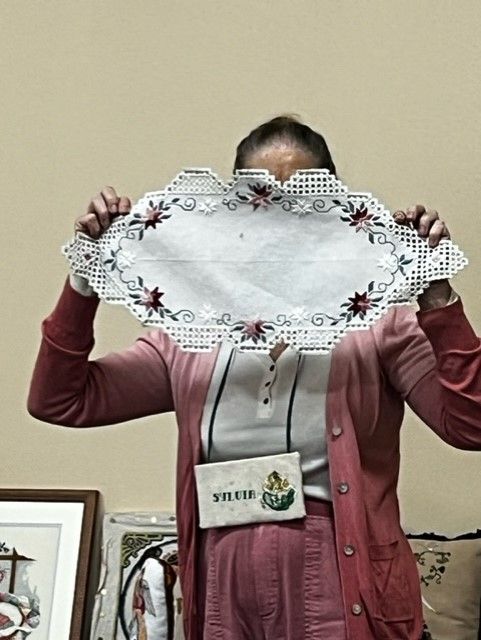 A person in a pink cardigan holds up an oval, white lace doily with floral embroidery in front of their face.