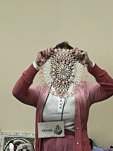 A person holds a round, intricate, cream-colored lace doily in front of their face against a plain, light-colored wall.