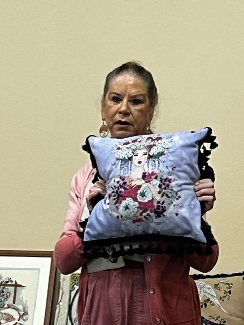 A person holds up a square, light-blue decorative throw pillow featuring an illustration of a face adorned with flowers.
