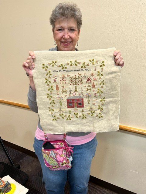A smiling person holds up a square piece of cream-colored fabric featuring a detailed, embroidered house and floral border.