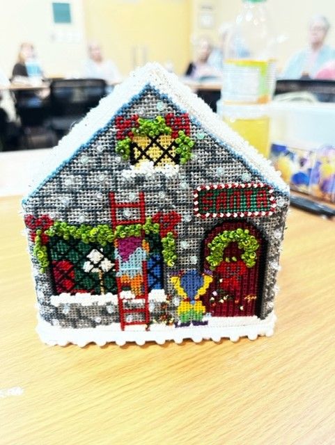 A handcrafted, cross-stitched 3D holiday house with a red ladder, an elf figure, and festive wreath decor on a wooden table.