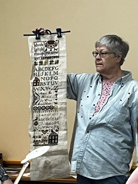 A person in a light blue shirt holds up a long piece of fabric featuring intricate black cross-stitched designs.
