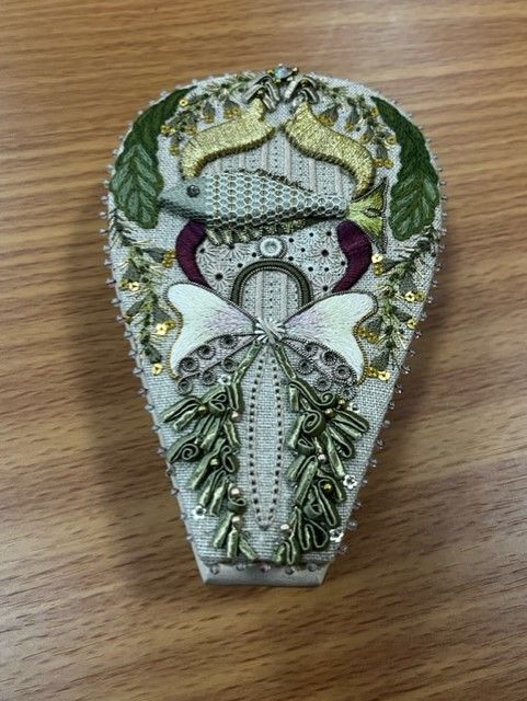 An intricate, pear-shaped embroidery piece featuring a central fish, a butterfly, green leaves, and gold accents.