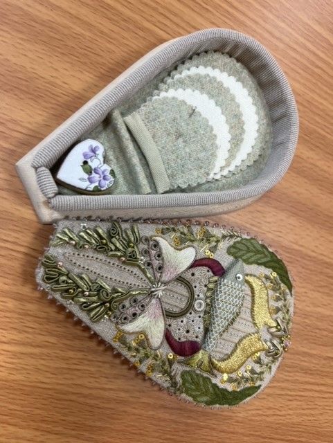 A decorated, teardrop-shaped case lying open on a wood surface, revealing fabric inserts and a small heart-shaped charm.