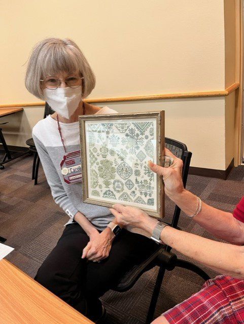 A person in a mask looks on as another person holds up a framed, light-colored square needlepoint art piece.