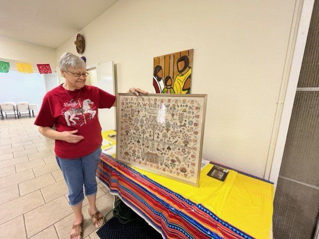 A person in a red shirt stands at a table, holding a framed piece of embroidery featuring many small, intricate designs.
