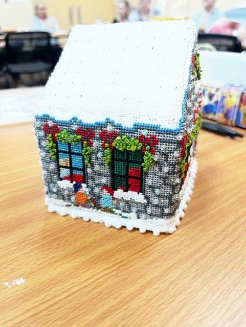 A decorative, small handcrafted house made of needlepoint or beads, featuring stone-textured walls, windows, and snow.