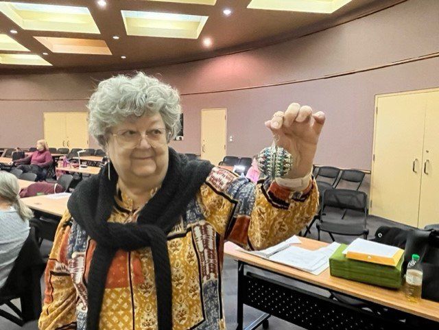 A person in a patterned shirt holds up a small, folded currency ornament in a room with rows of empty chairs.