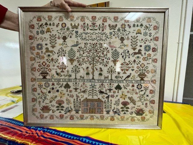 A framed, detailed sampler featuring colorful cross-stitch patterns of a house, animals, and flowers with embroidered text.