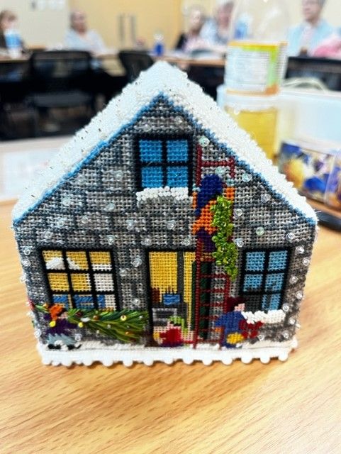 A 3D needlepoint house featuring gray stone walls, snowy roof, decorated windows, a ladder, and people carrying items.