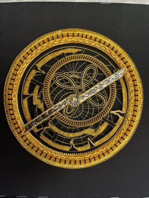 A circular, intricate gold astrolabe featuring filigree patterns and a silver-toned arm set against a dark background.