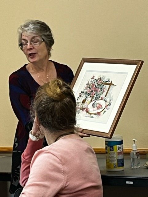 A woman with glasses displays a framed floral artwork to a person with hair in a bun in a bright, indoor room.