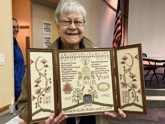 A smiling person holds an open, triptych-style embroidery piece featuring flowers, text, a house, and the year 2022.