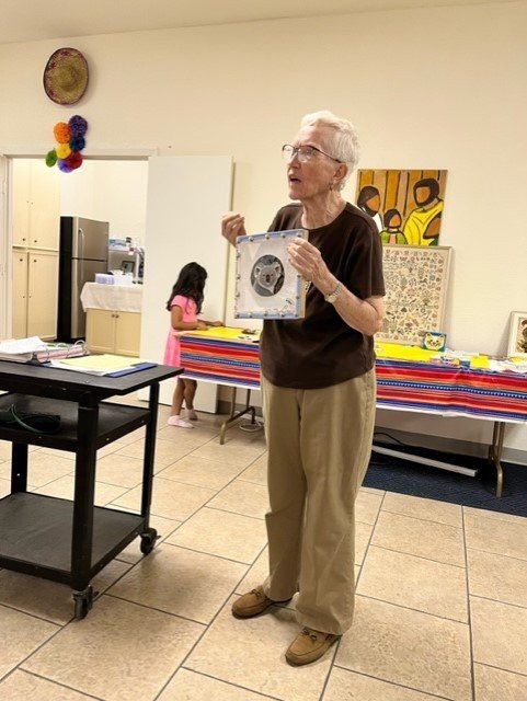 A person stands in a room holding up a small, framed square object while a child stands at a table in the background.