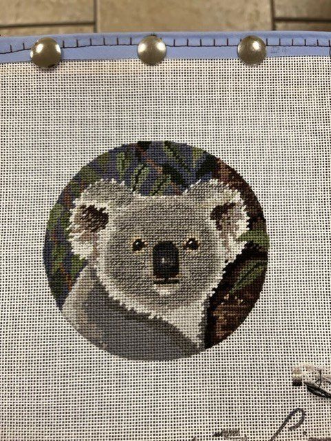 A needlepoint embroidery of a grey koala centered in a circular design against a dark, leafy background.