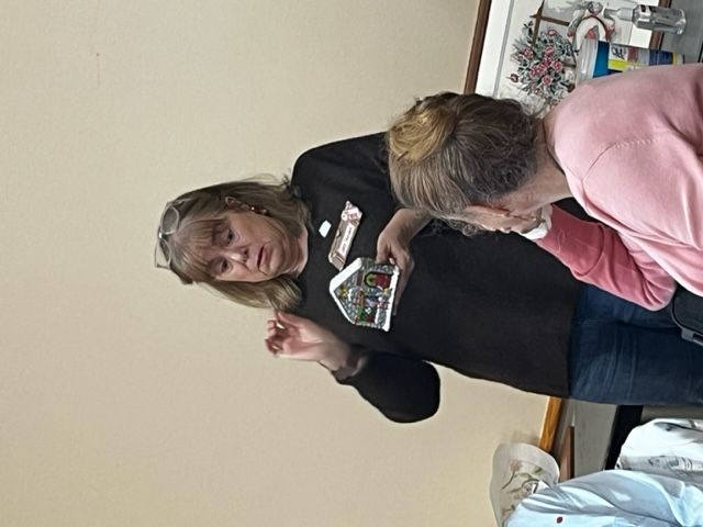 A person wearing glasses and a dark shirt holds up a small, decorated model house while speaking to a group.