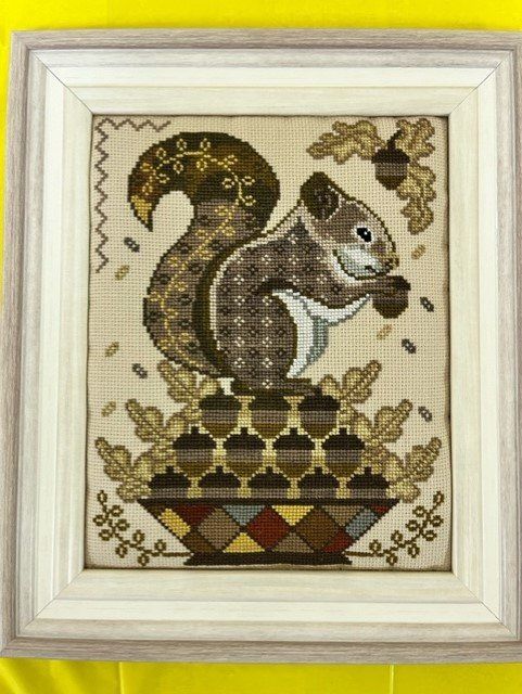 A cross-stitch of a brown squirrel holding an acorn, sitting atop a decorative basket of acorns against an oak leaf motif.