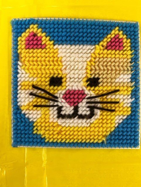A yellow and white needlepoint cat face on a blue background with pink inner ears and a pink nose.