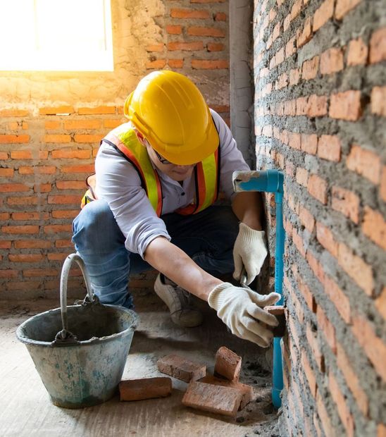 A man wearing a yellow hard hat and safety vest is working on a brick wall.