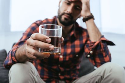 Man sitting on the floor, holding a glass of whiskey and looking stressed with his hand on his forehead