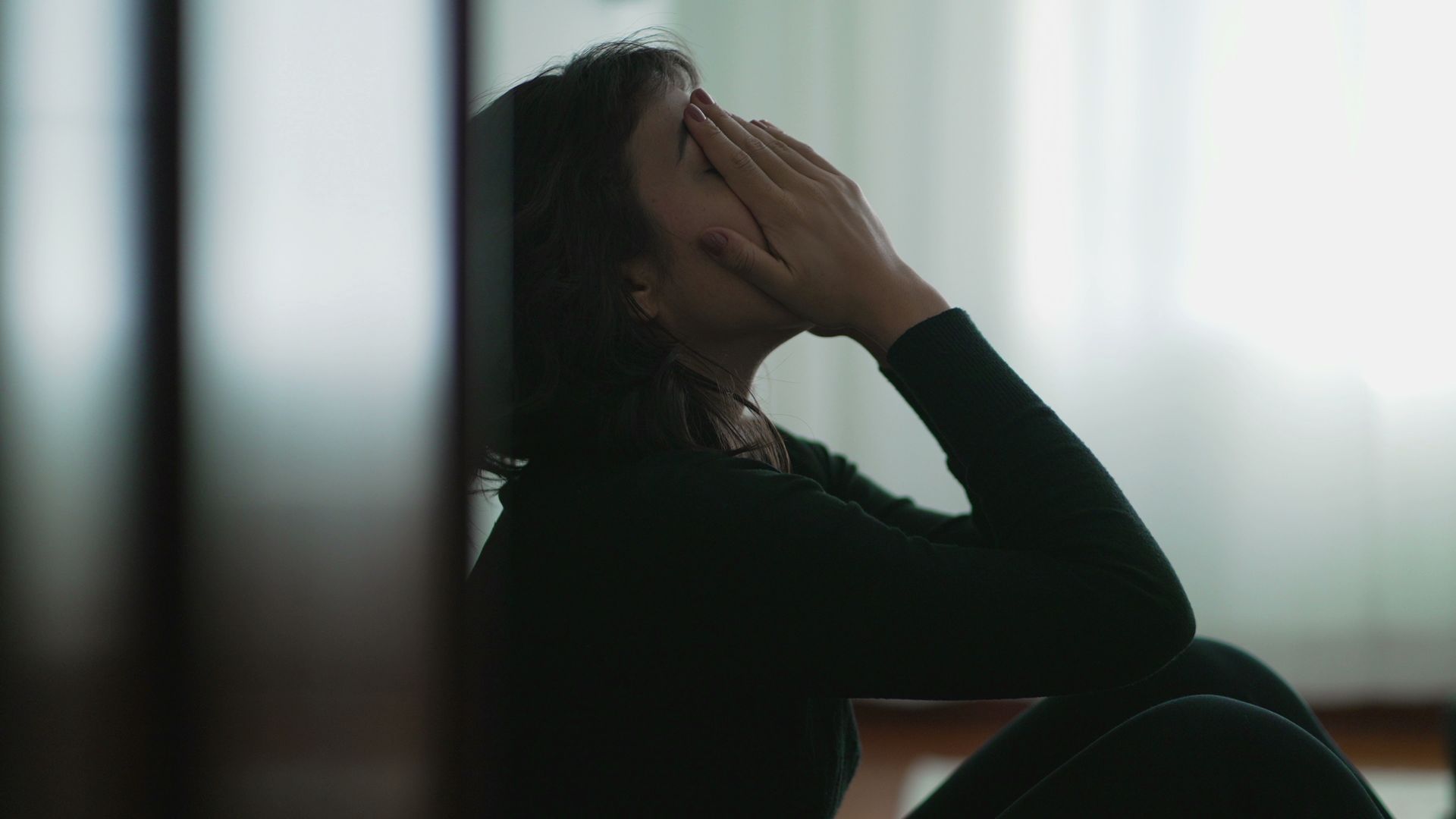 Person sitting by a window, head in hand, silhouetted against soft daylight.