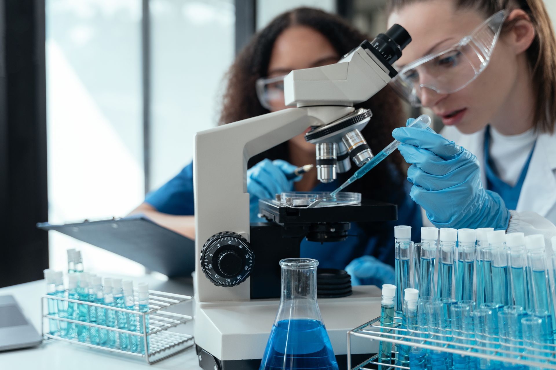Scientists using a microscope in a lab, with blue-gloved hands and test tubes nearby