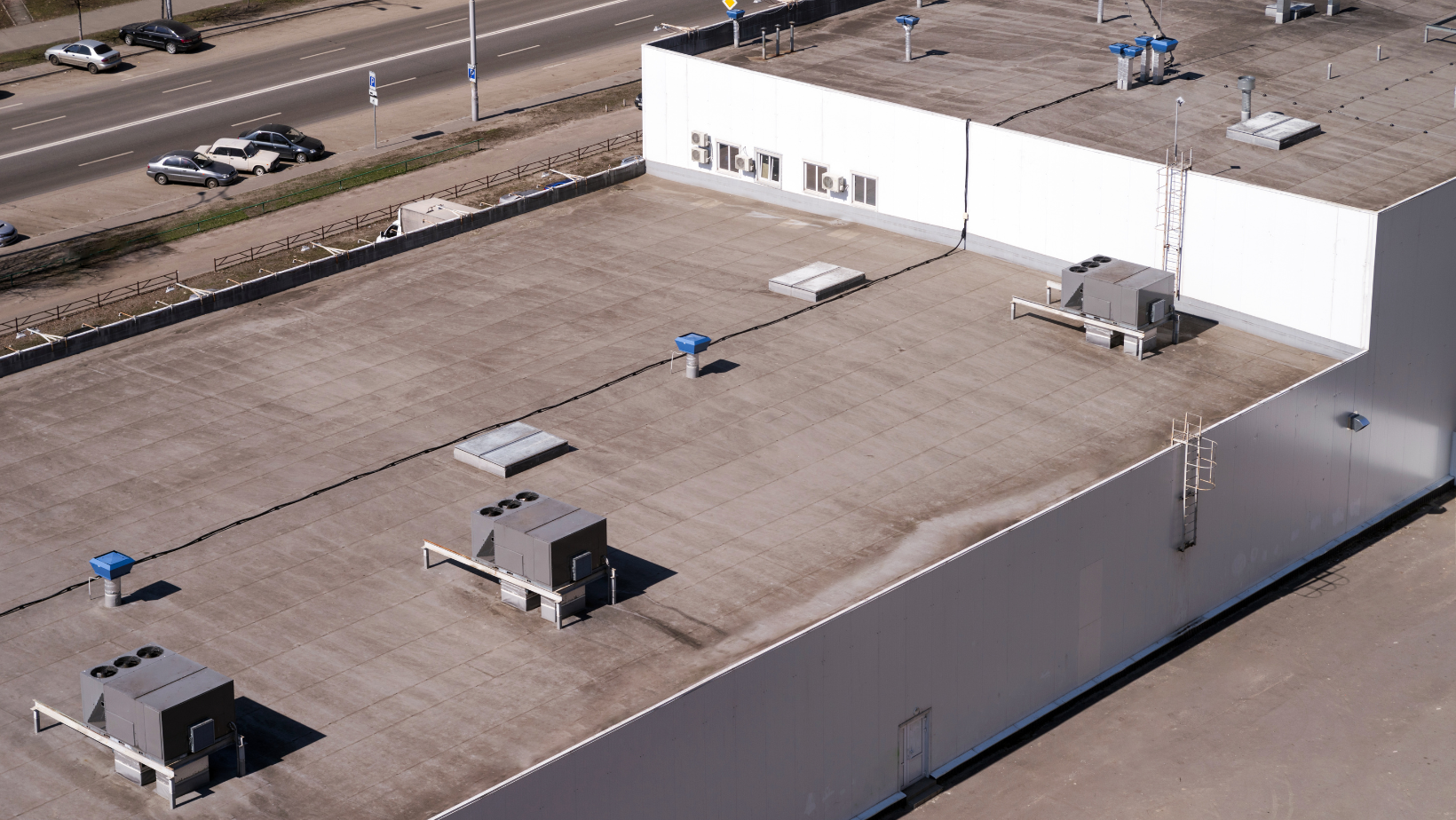 Aerial view of a large flat-roofed warehouse with HVAC units and a white exterior wall along a roadway