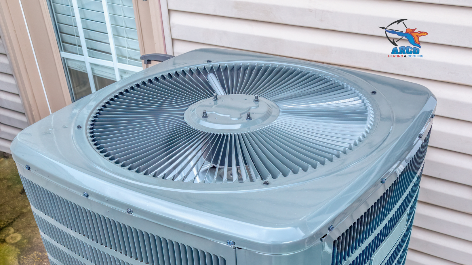 Gray air conditioning unit outside a building with white siding. Fan on top.