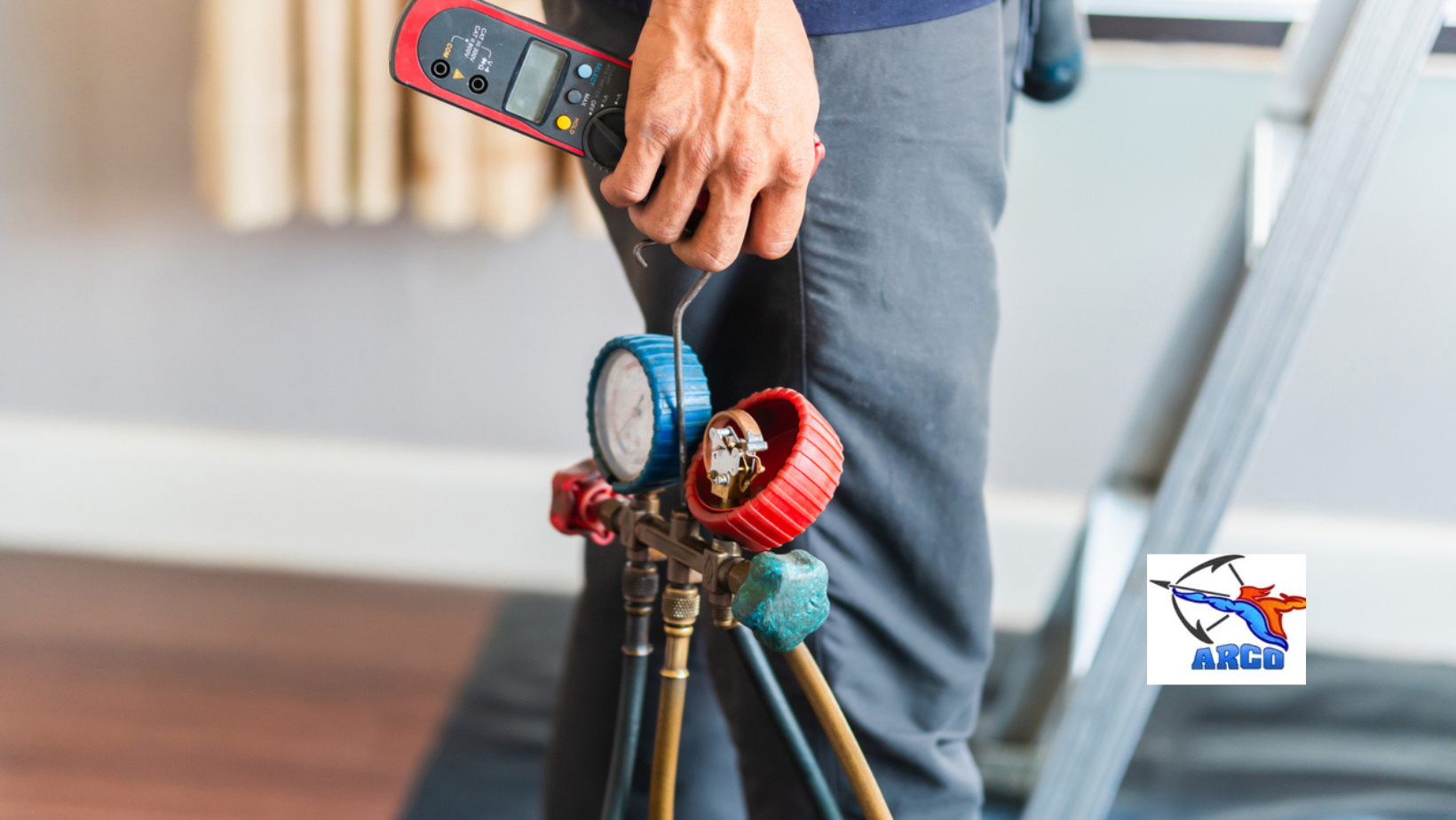 ARCO HVAC technician holding gauges and a digital meter near a ladder indoors.