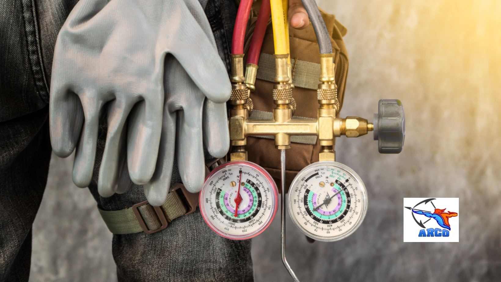 Gloved worker holding HVAC manifold gauges with red and yellow hoses attached to brass valves