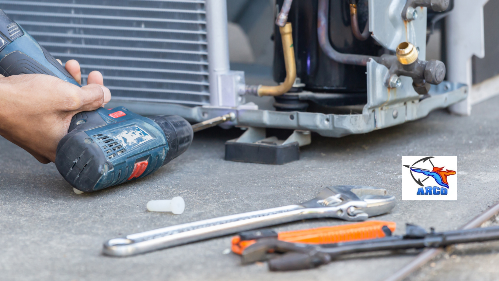 Hand using a blue cordless drill near an outdoor HVAC unit, with tools on the ground.