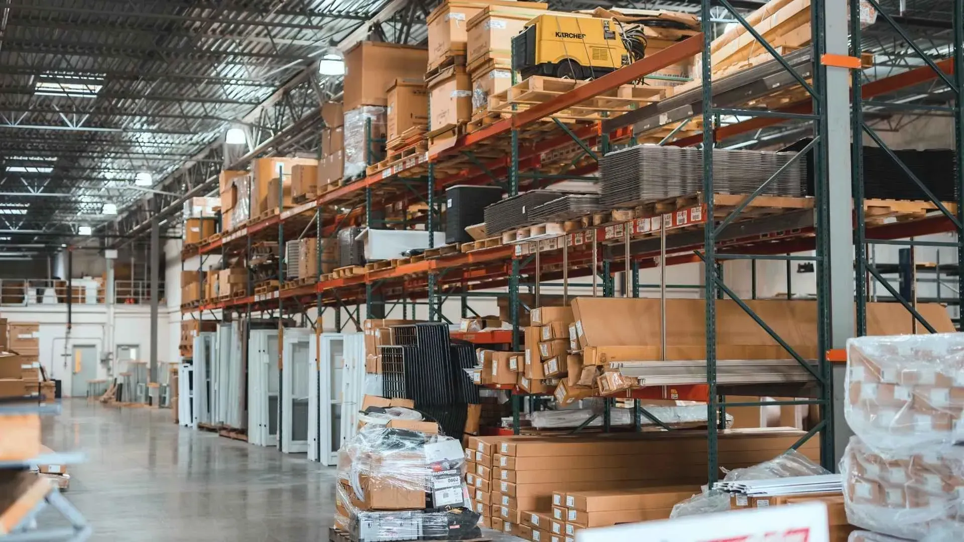 Warehouse aisle with tall orange shelving, stacked boxes, and a worker using a forklift