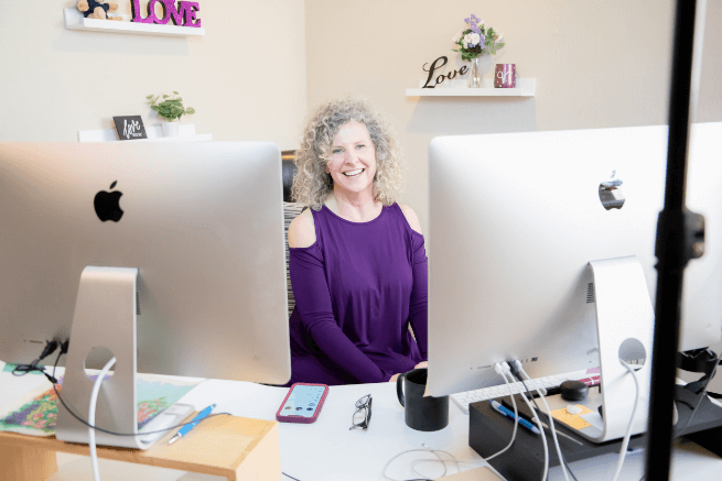 Woman seated between two Apple computers, smiling. She wears a purple cold-shoulder top in an office setting.