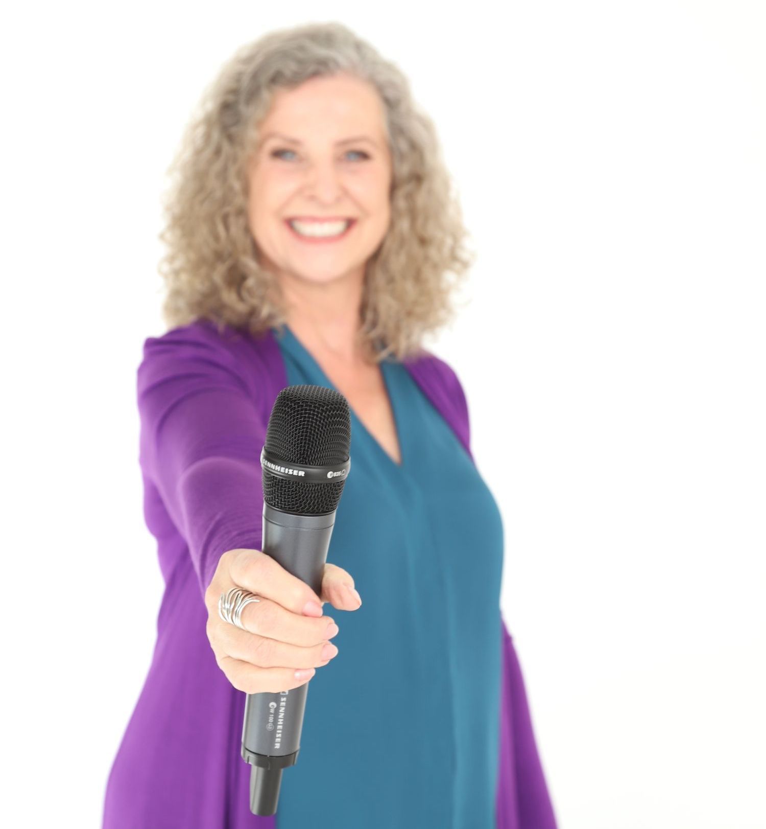 Woman holding out microphone, smiling, wearing teal top and purple cardigan.