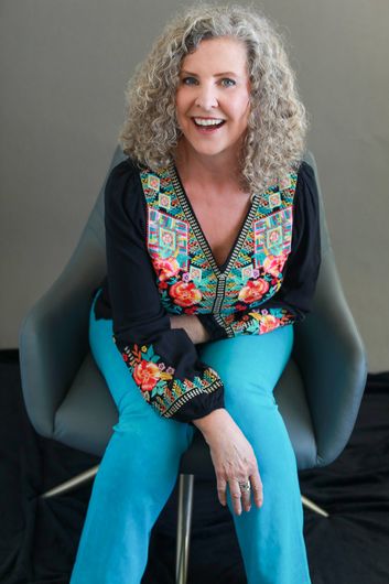 Woman with curly grey hair smiles, seated in a chair, wearing a floral embroidered top and teal pants.