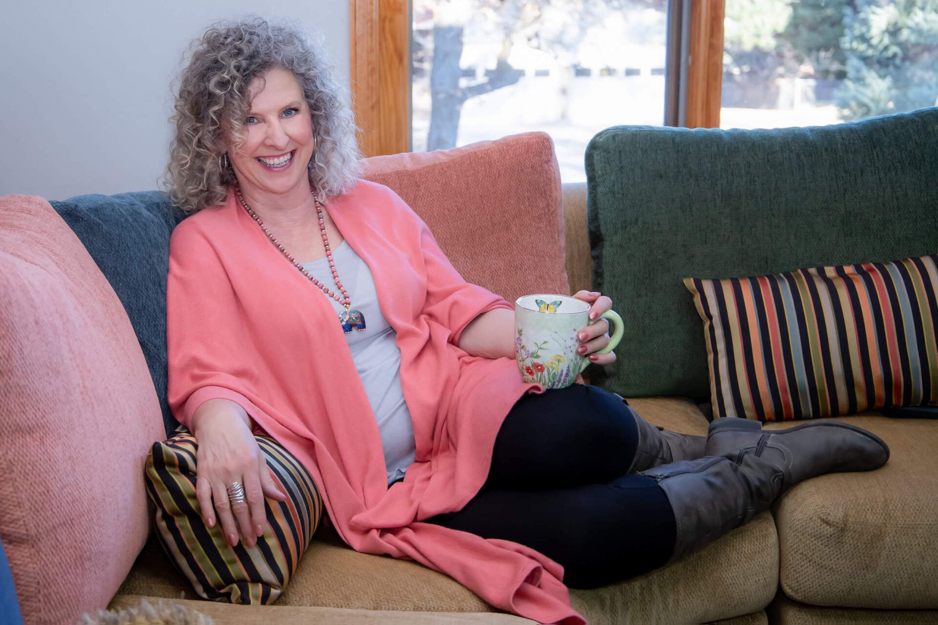 Woman lounging on a couch, holding mug, wearing pink cardigan, smiling, near window with colorful pillows.