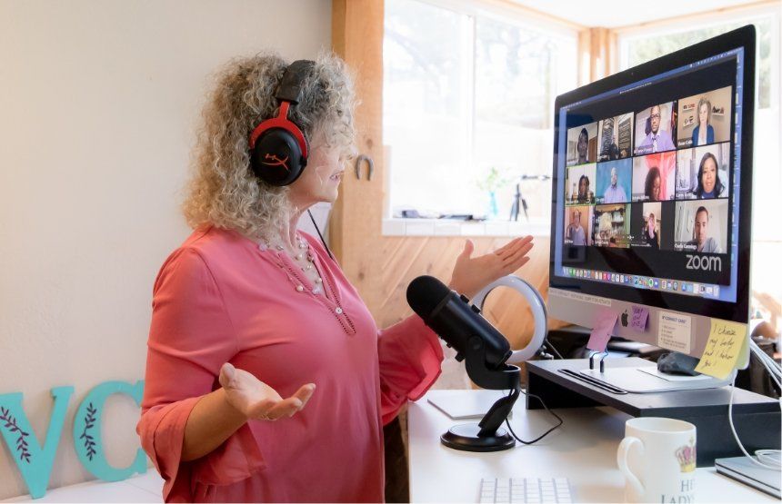 Woman wearing headphones speaks during video call, gesturing with hands. A computer screen displays other participants.