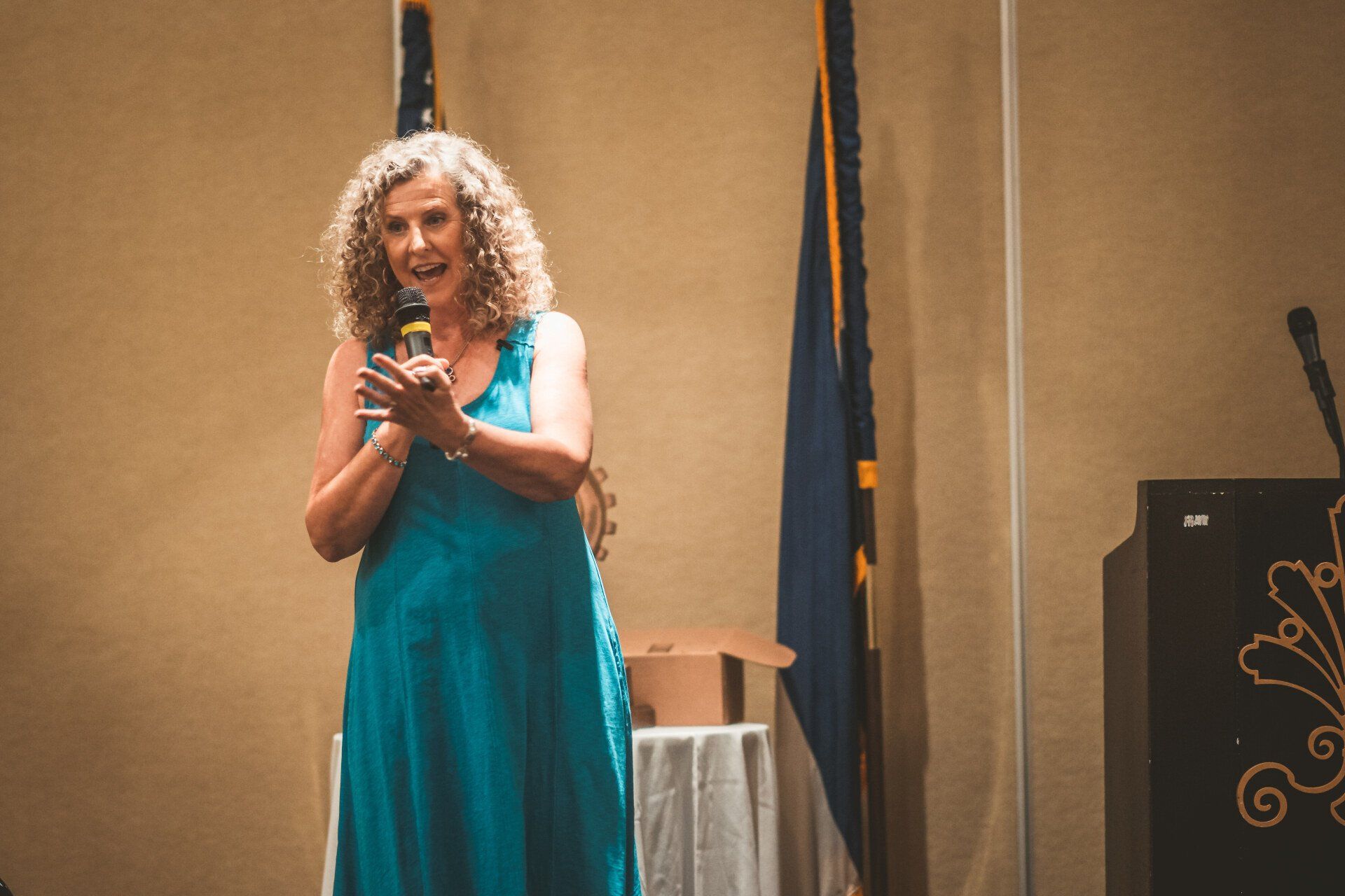 Woman in a teal dress speaks into a microphone, clapping her hands on a stage with flags behind her.