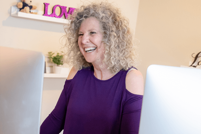 Woman with curly gray hair smiles while looking at computer screens in a home office with a 