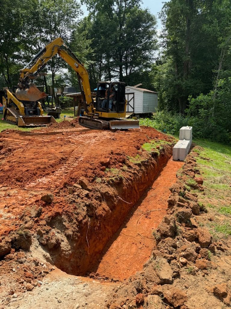 Excavator digging a long trench in red clay beside a wooded area and shed