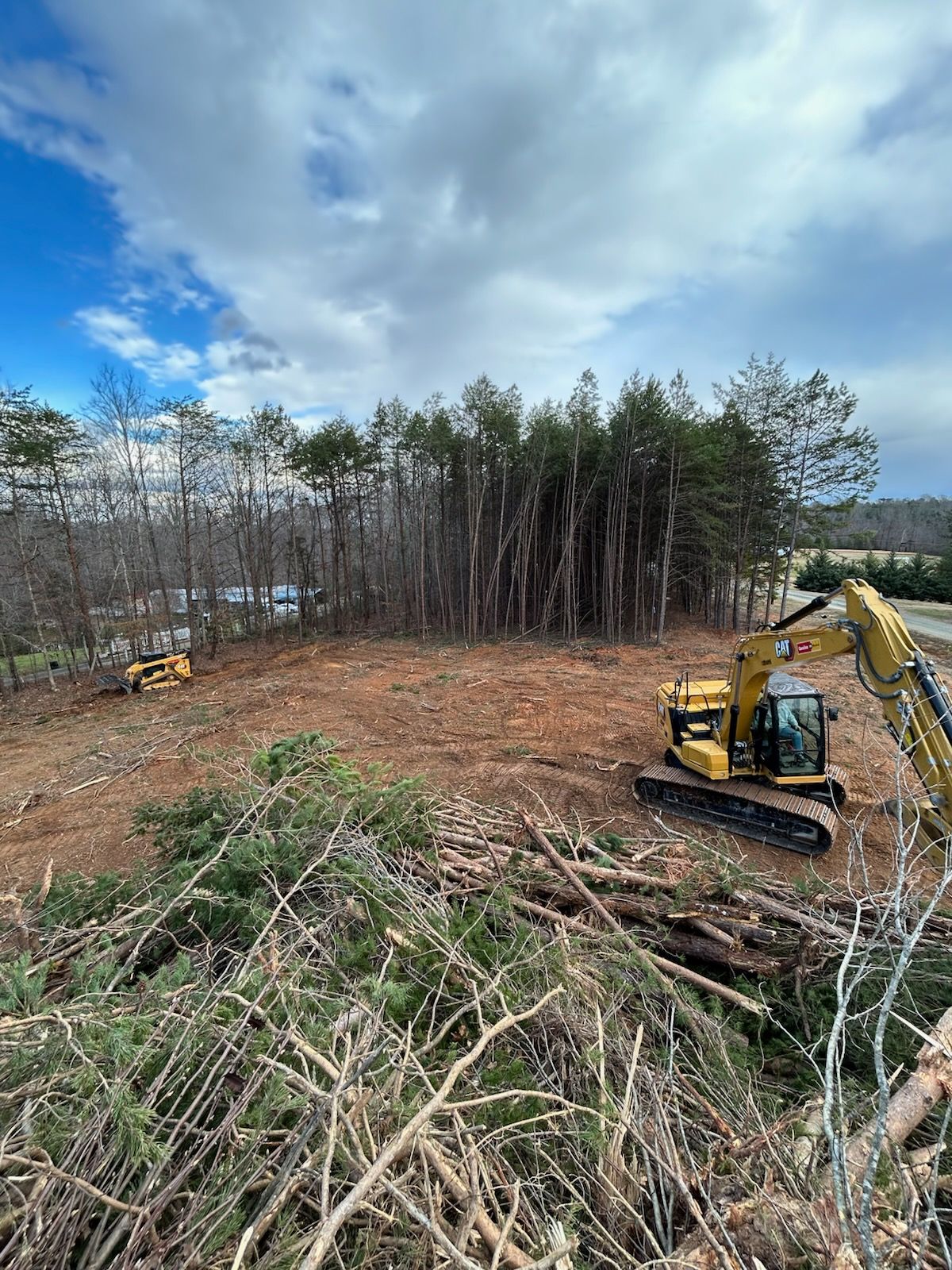 Excavator clearing a wooded lot under a cloudy sky, with cut trees and brush piled in the foreground
