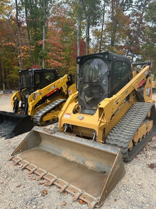 Two yellow skid steer loaders with buckets parked on gravel in a wooded area