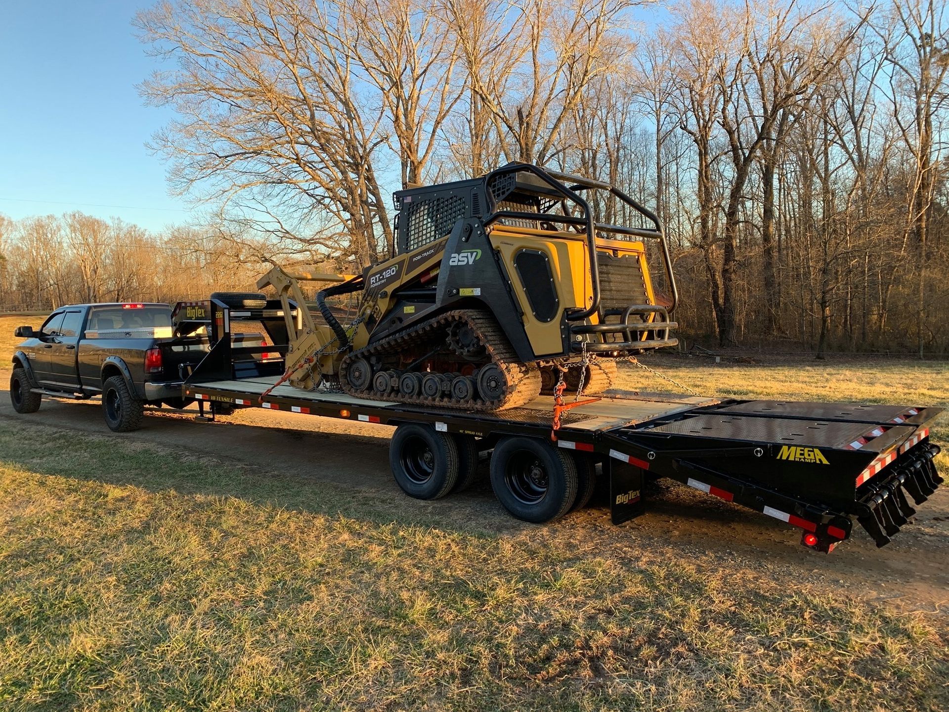 Pickup truck towing a skid-steer loader on a flatbed trailer beside leafless trees at dusk