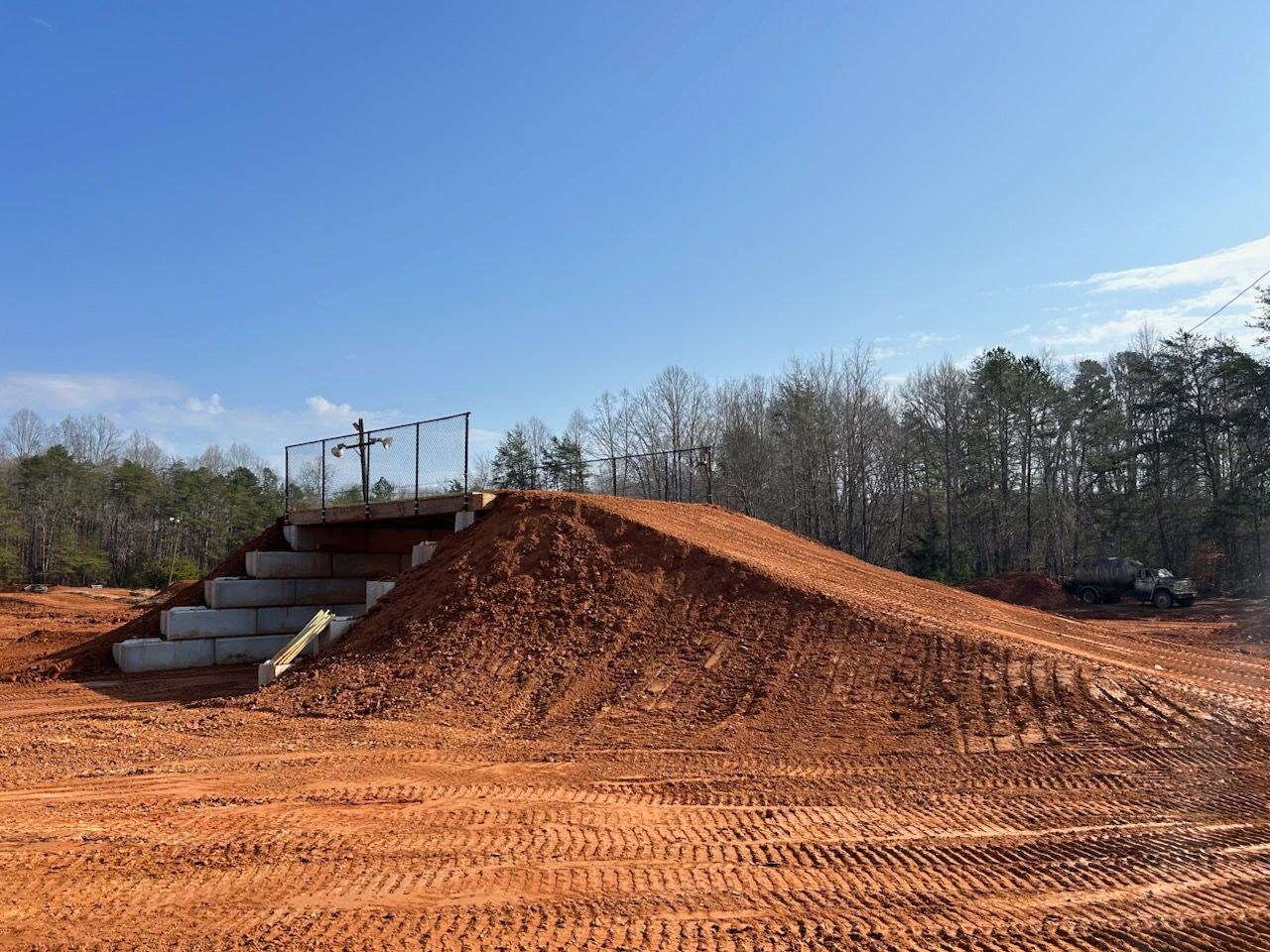 Dirt mound with a small concrete stairway and railing under a clear blue sky, in a wooded area.