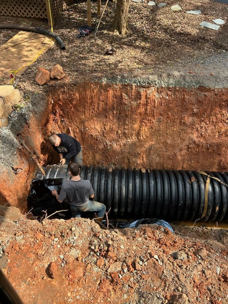 Workers installing a black drainage pipe in a dug trench outdoors.