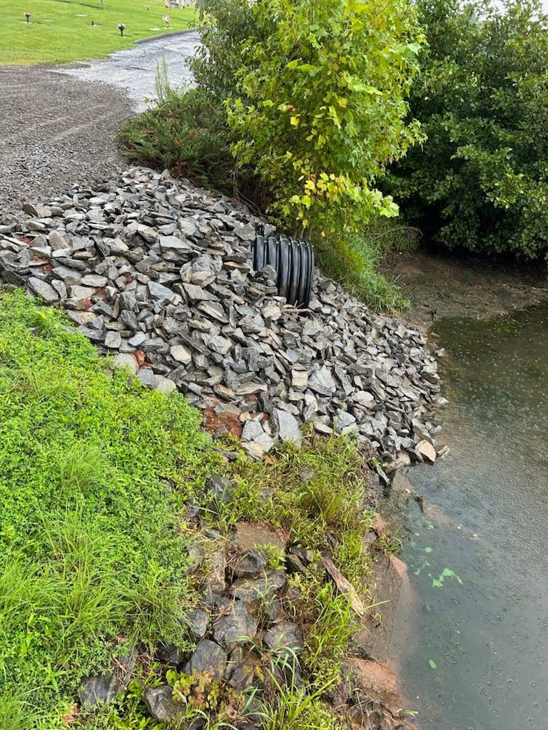 Rock-lined creek bank with a black drainage pipe beside green grass and water