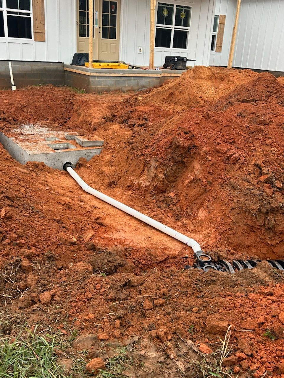 Reddish dirt excavation in front of a house, with a white pipe and exposed drain cover.
