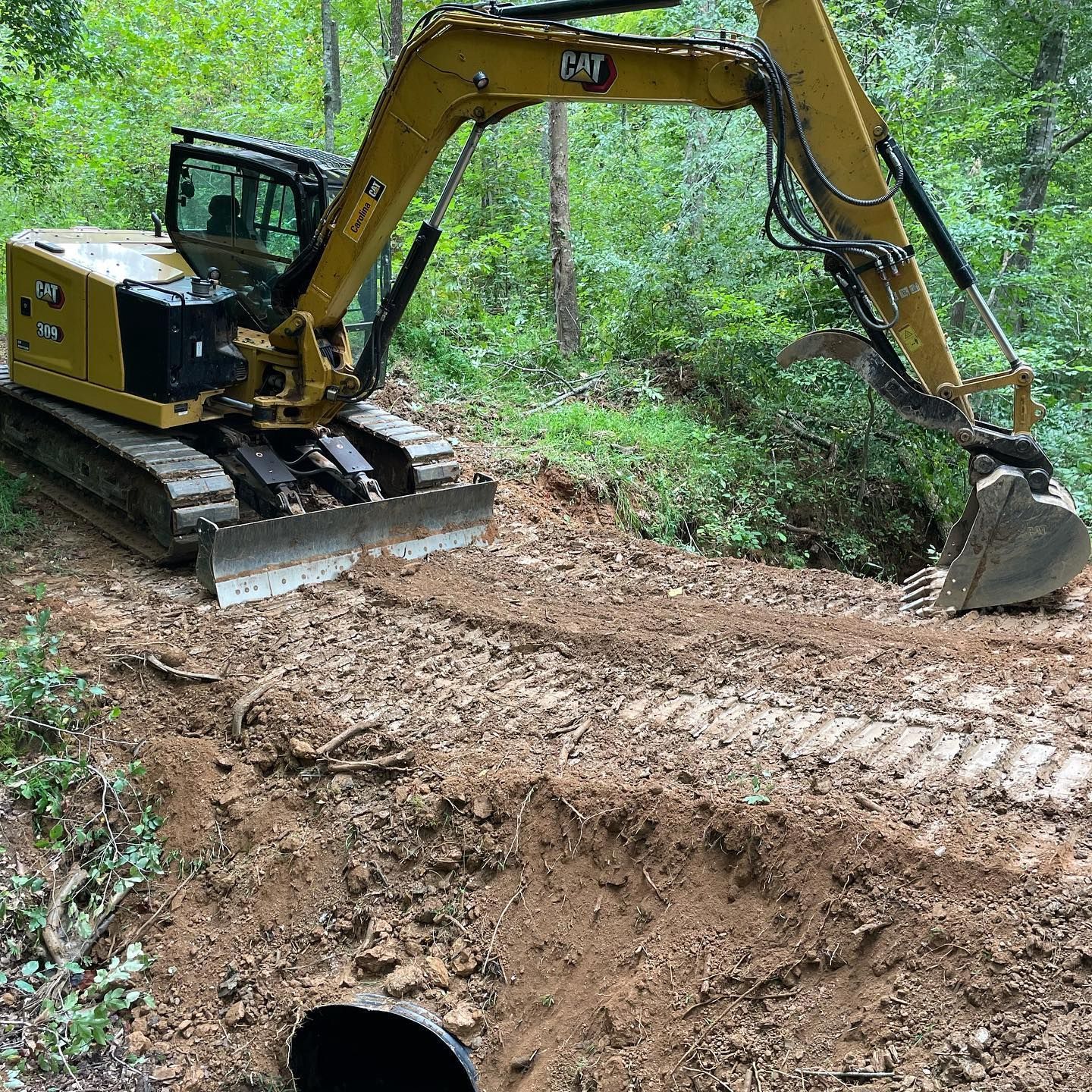 Yellow excavator digging a muddy trench beside a black pipe in a wooded area