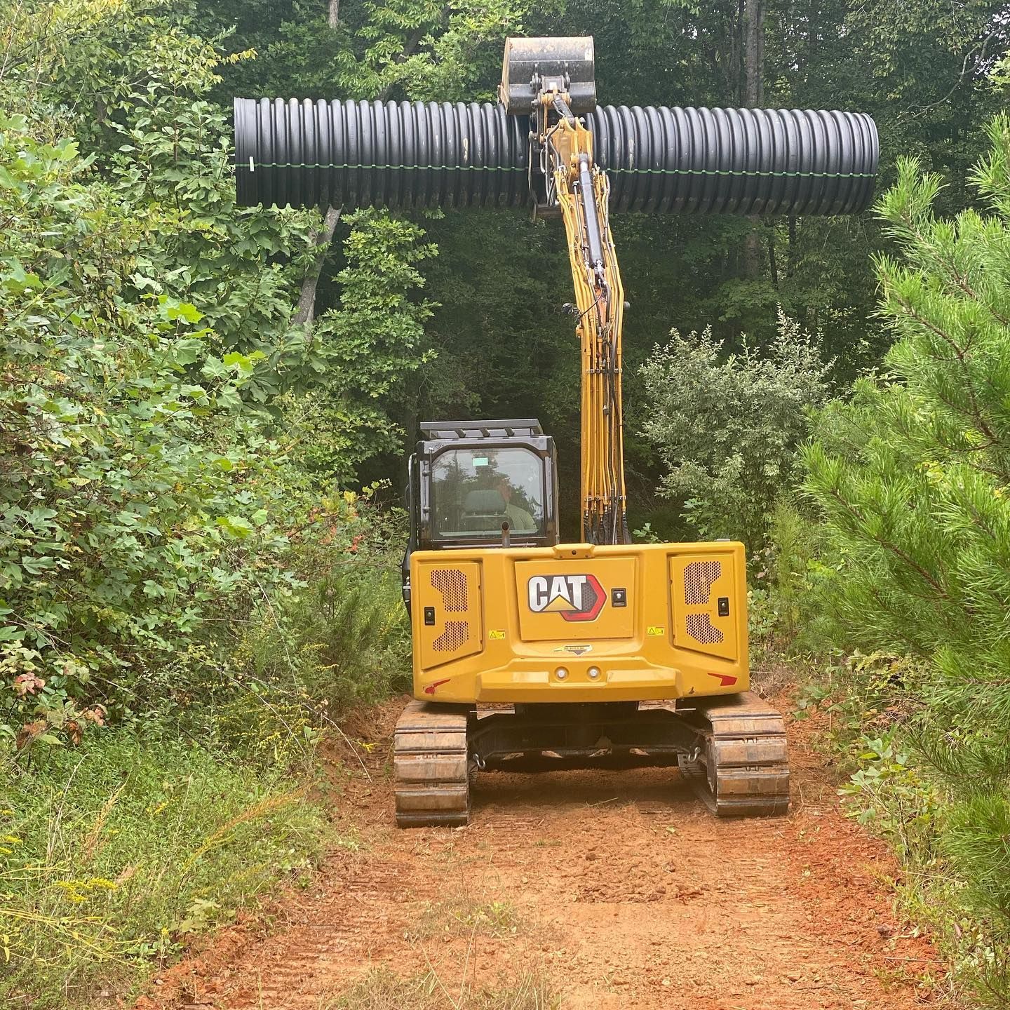 Yellow CAT excavator with a long black attachment clearing a muddy path in dense green woods
