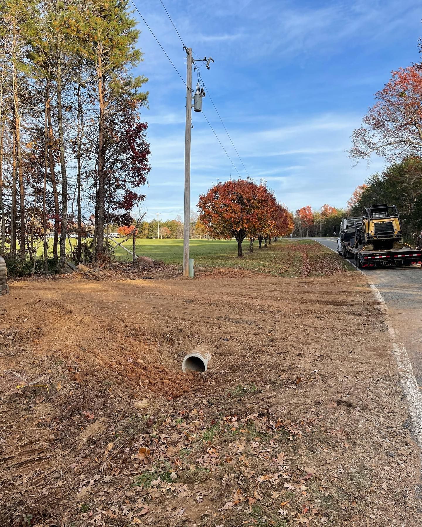 Rural roadside with autumn trees, utility pole, drainage culvert, and construction equipment beside the road
